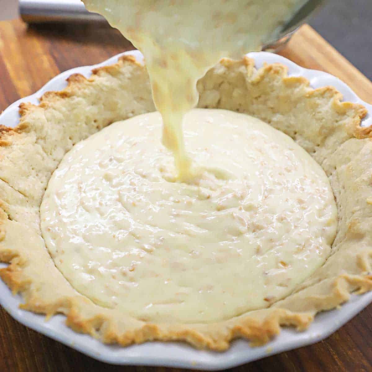 Coconut cream pie filling being poured into a baked pie crust in a white pie dish. 