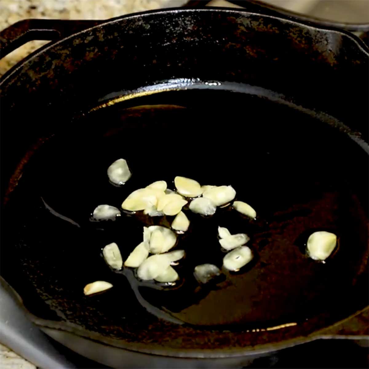 Slices of garlic being sautéed in olive oil in a large cast-iron skillet. 