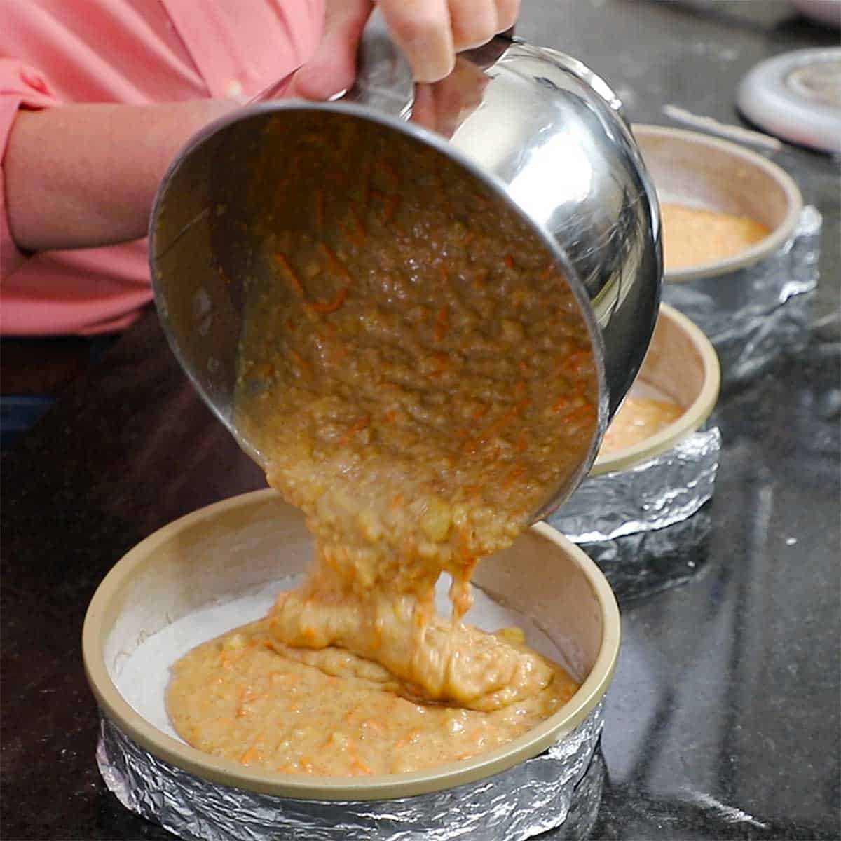 A person pouring carrot cake batter into a cake pan from a large silver mixing bowl. 