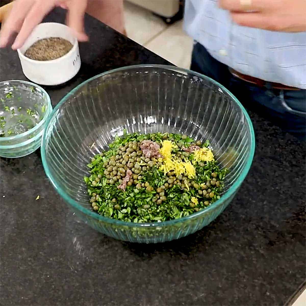 A glass bowl filled with chopped fresh herbs, garlic, pepper, lemon zest, and anchovies. 