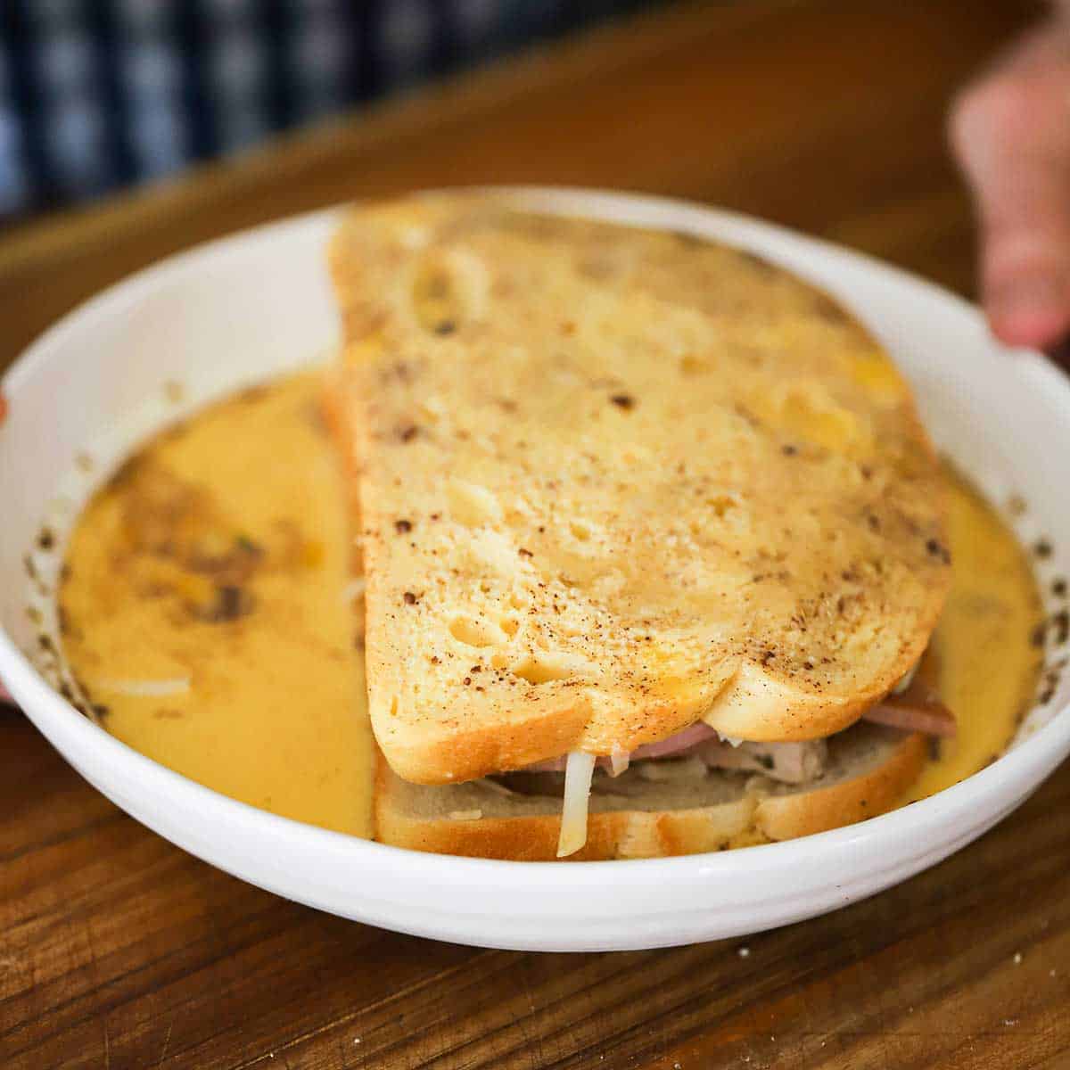 A person holding onto a white bowl that is filled with a ham, turkey, and cheese sandwich that has been submerged on both sides in a cinnamon egg mixture. 