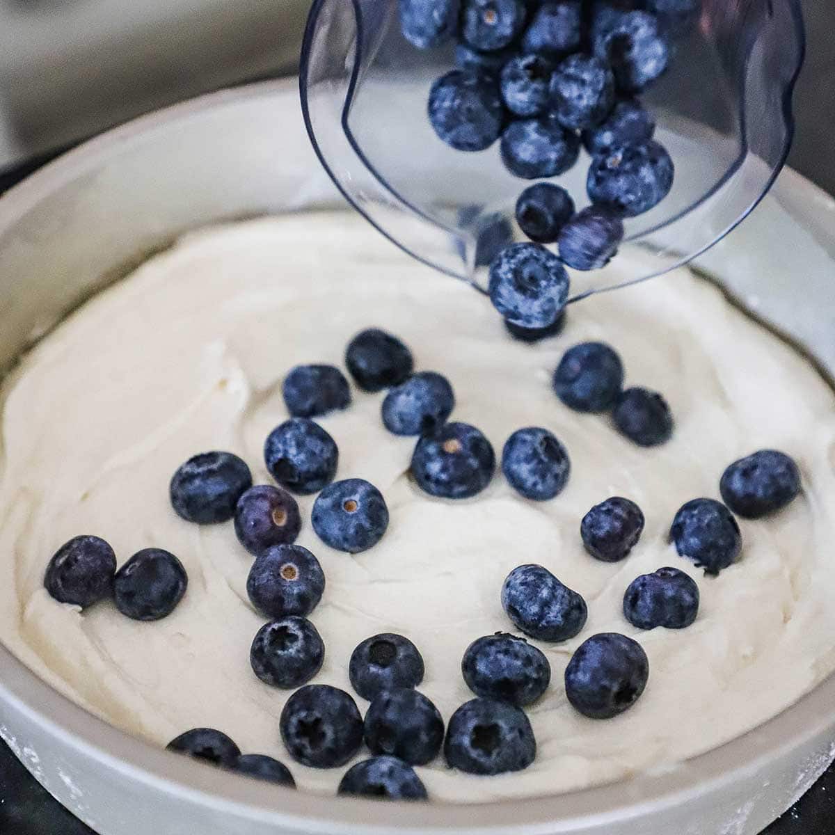 A person sprinkling blueberries from a glass bowl onto the top of coffee cake batter that is resting in a metal cake pan. 