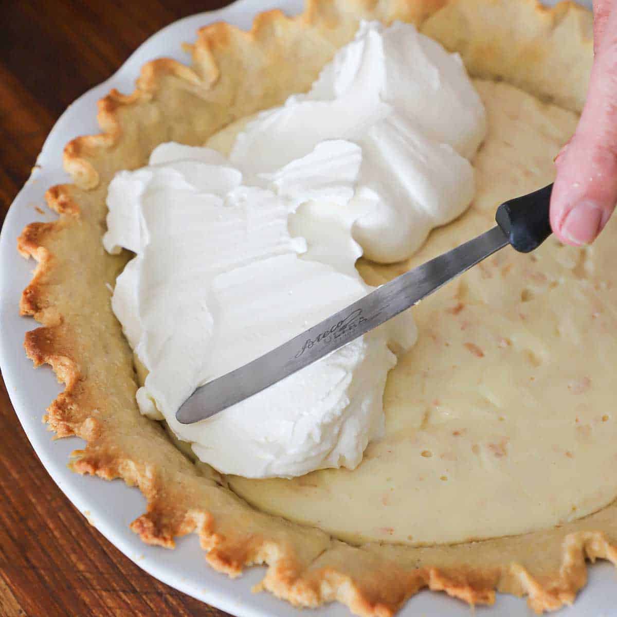 A person using an offset spatula to spread whipped cream over the top of a chilled coconut cream pie. 