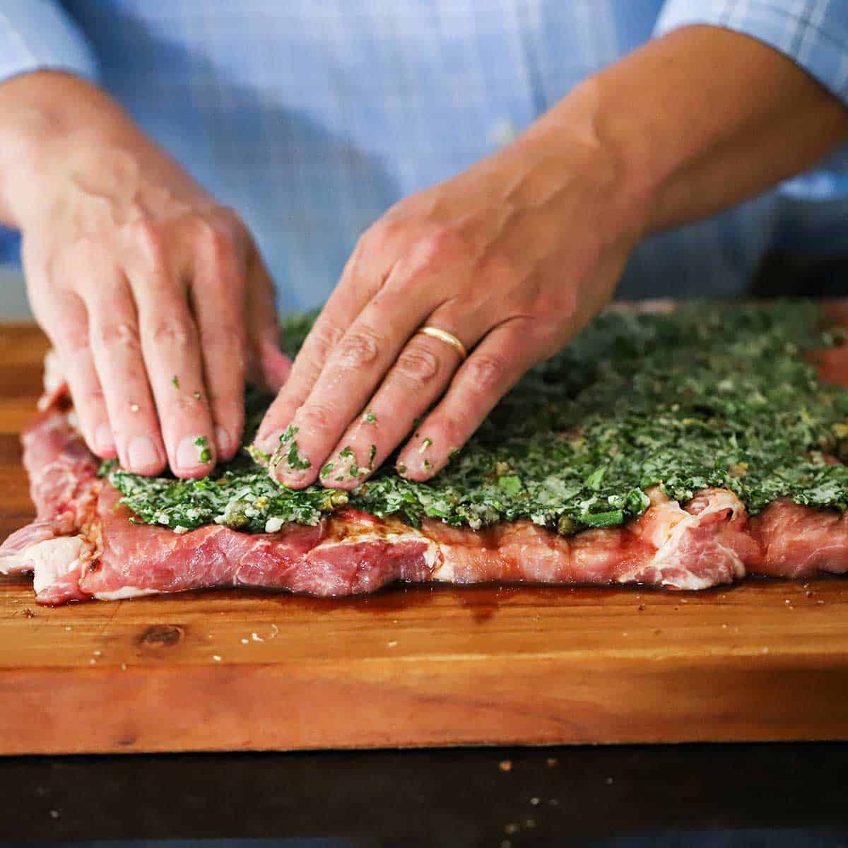 A person using his hands to spread an herb and garlic mixture of the surface of a pork loin that has been butterflied and opened up flat. 