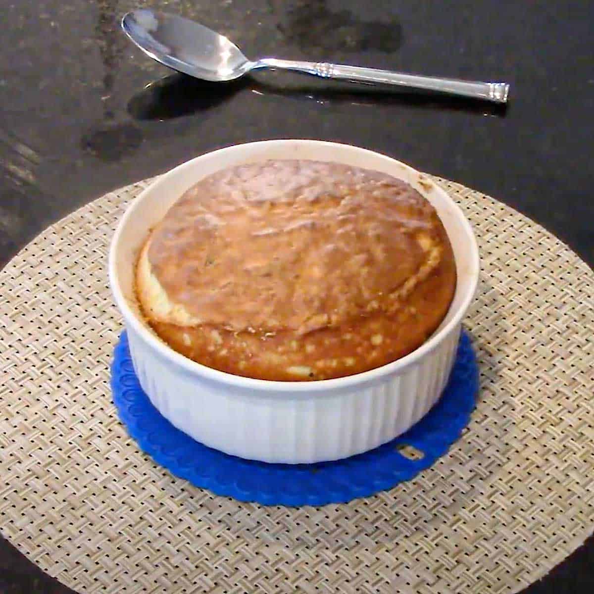 A fully baked cheese soufflé with a browned surface sitting on a blue hot pad on a circular woven place mat.