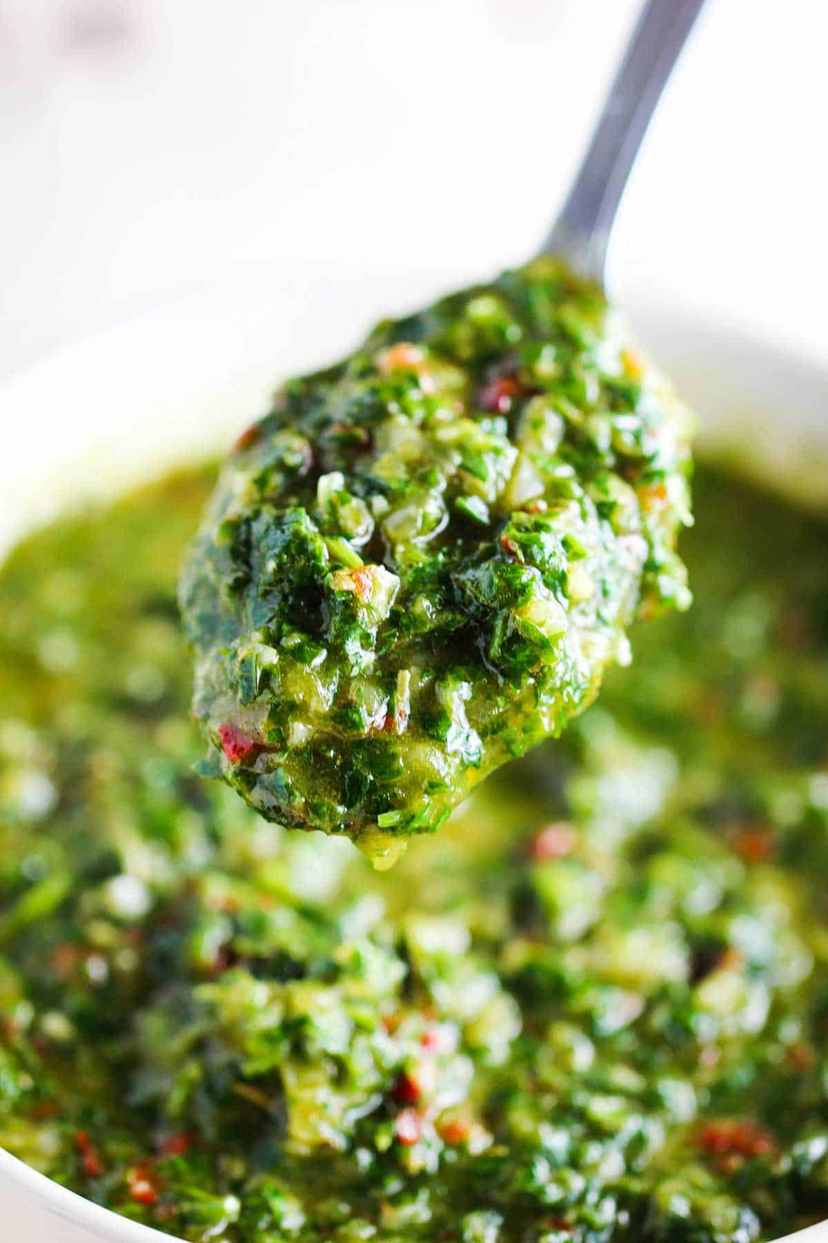 A spoon holding up a helping up authentic chimichurri sauce over a bowl of the same.