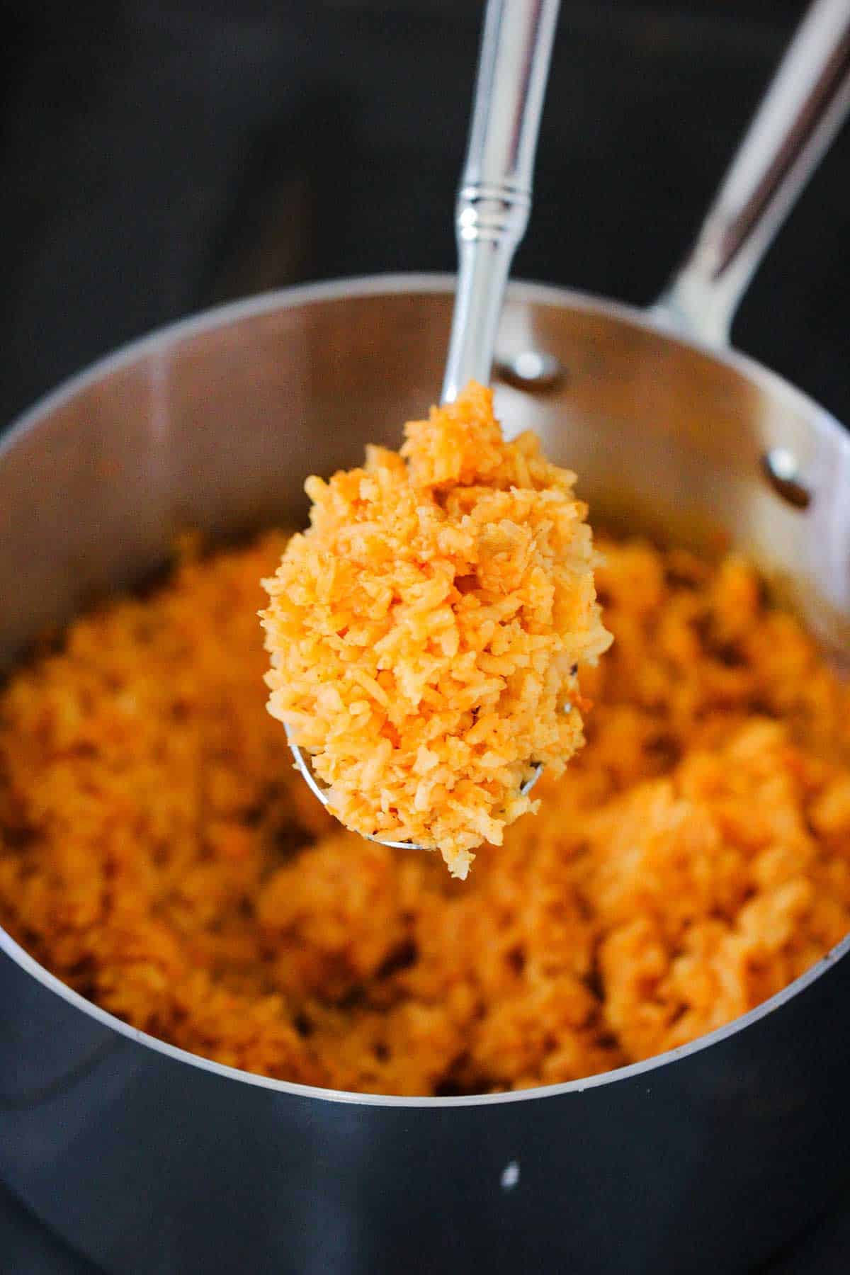 A person holding up a large spoon of homemade Mexican rice over a silver saucepan of the same.