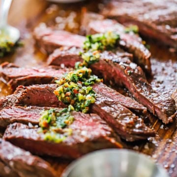 Strips of grilled carne asada that has been cooked to medium-rare and is resting on a cutting board and topped with chimichurri sauce and coarse sea salt.