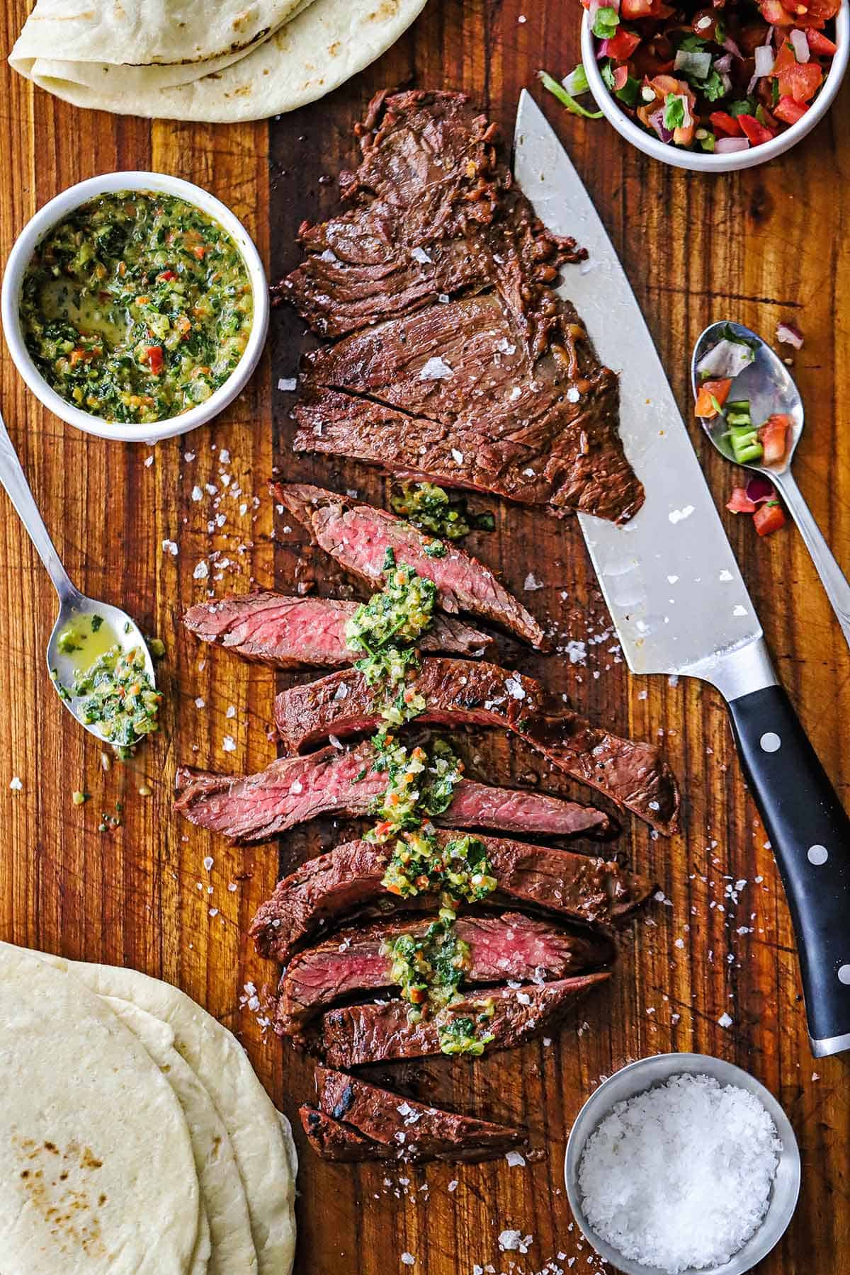 A partially sliced grilled carne asada on a wooden cutting board topped with homemade chimichurri sauce with a large chef's knife near the meat along with tortillas, and a bowl of salt, and a bowl of pico de gallo.
