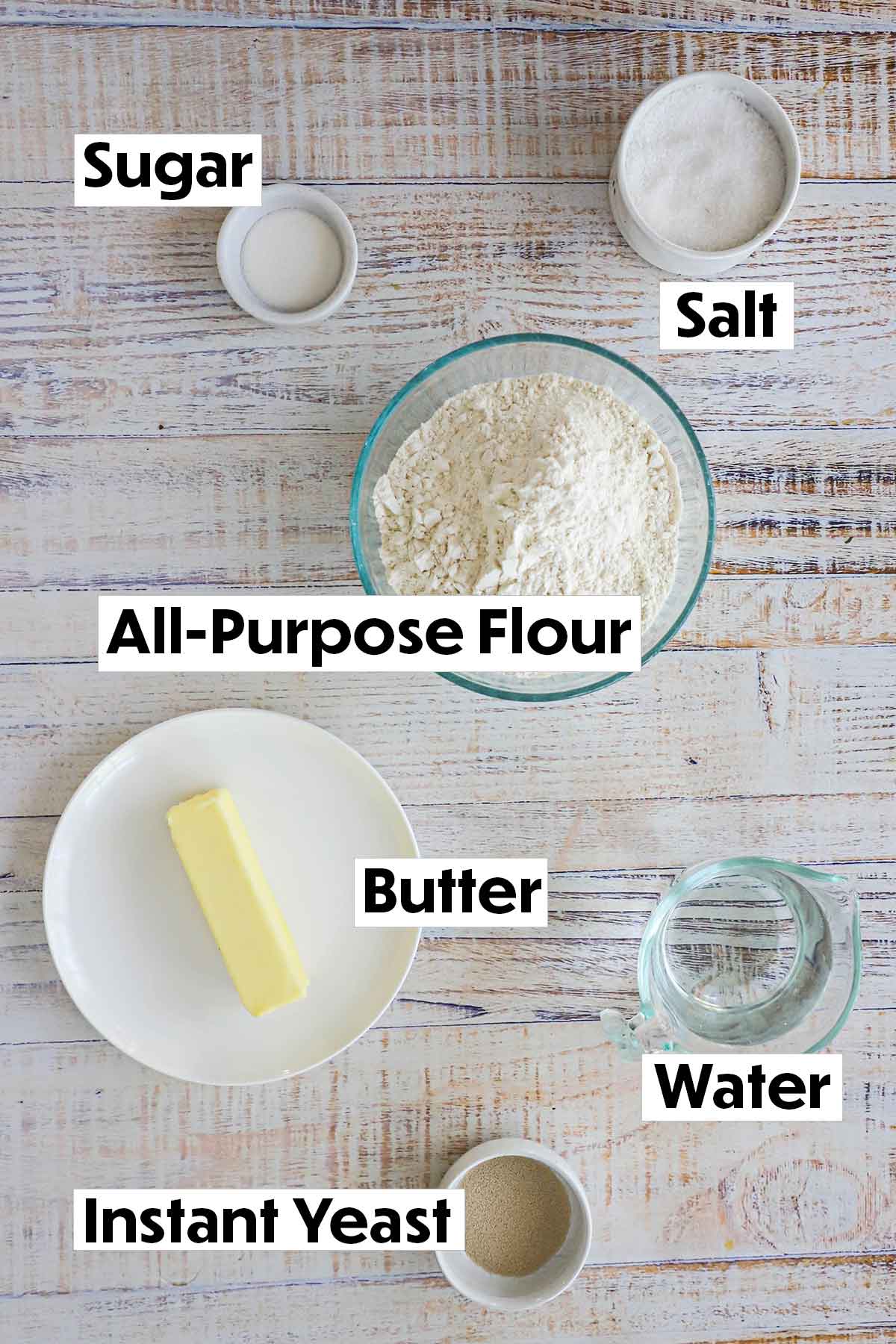 An arrangement of ingredients for country white bread on a white background including flour, sugar, salt, a stick of butter, water, and a small white bowl filled with instant yeast.