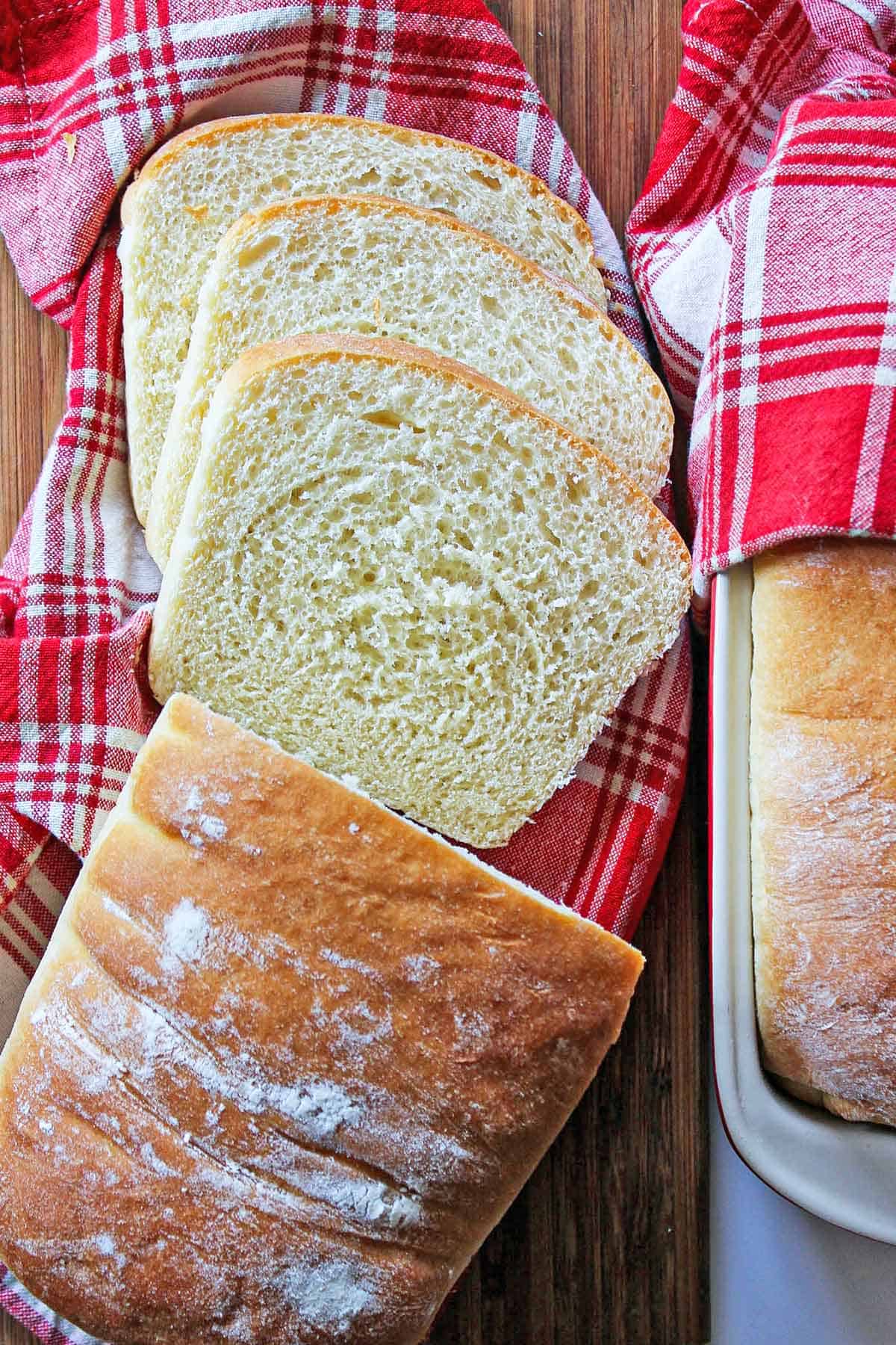A red and white plaid napkin with several slices of county white bread on it next to the remaining unsliced loaf.
