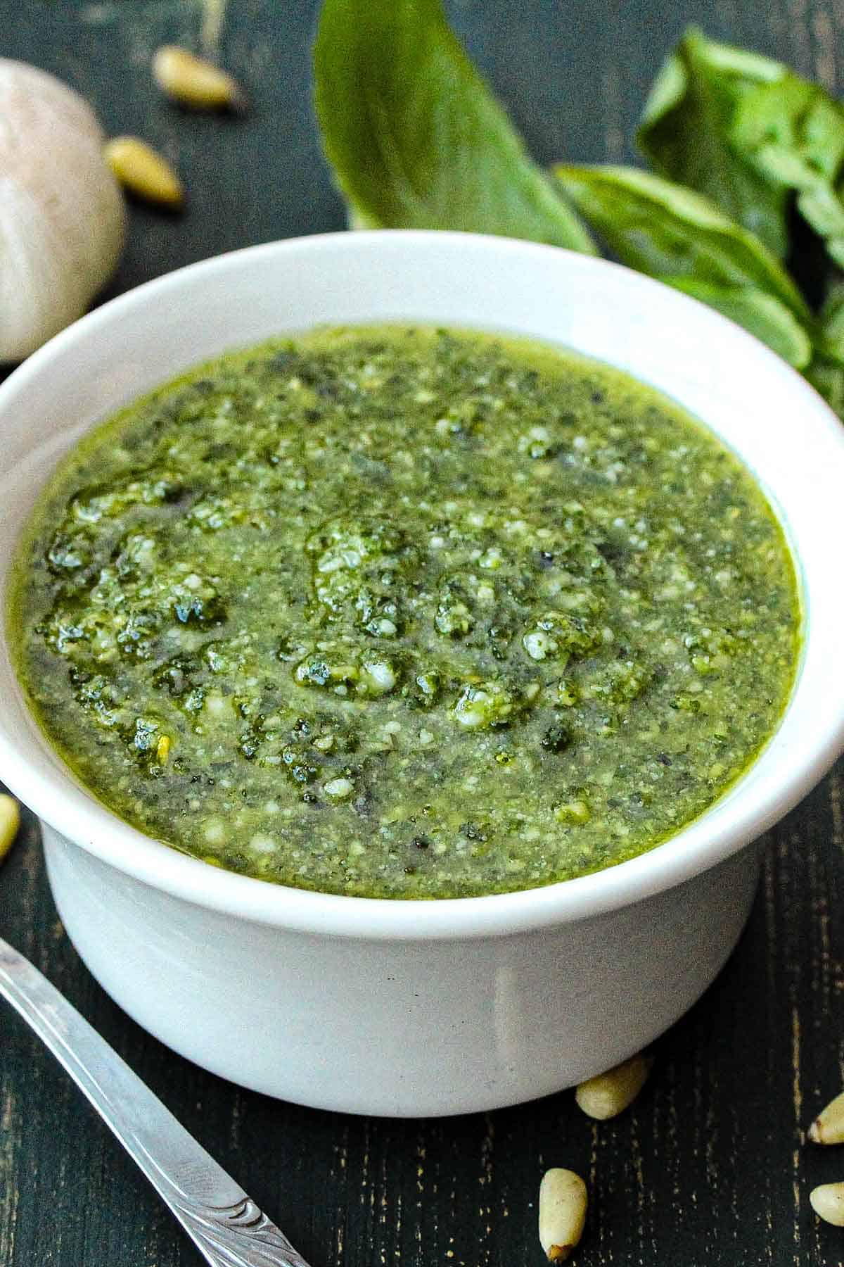 A bowl of easy pesto sauce in a small white bowl next to scattered pine nuts, a head of garlic, and fresh basil leaves.