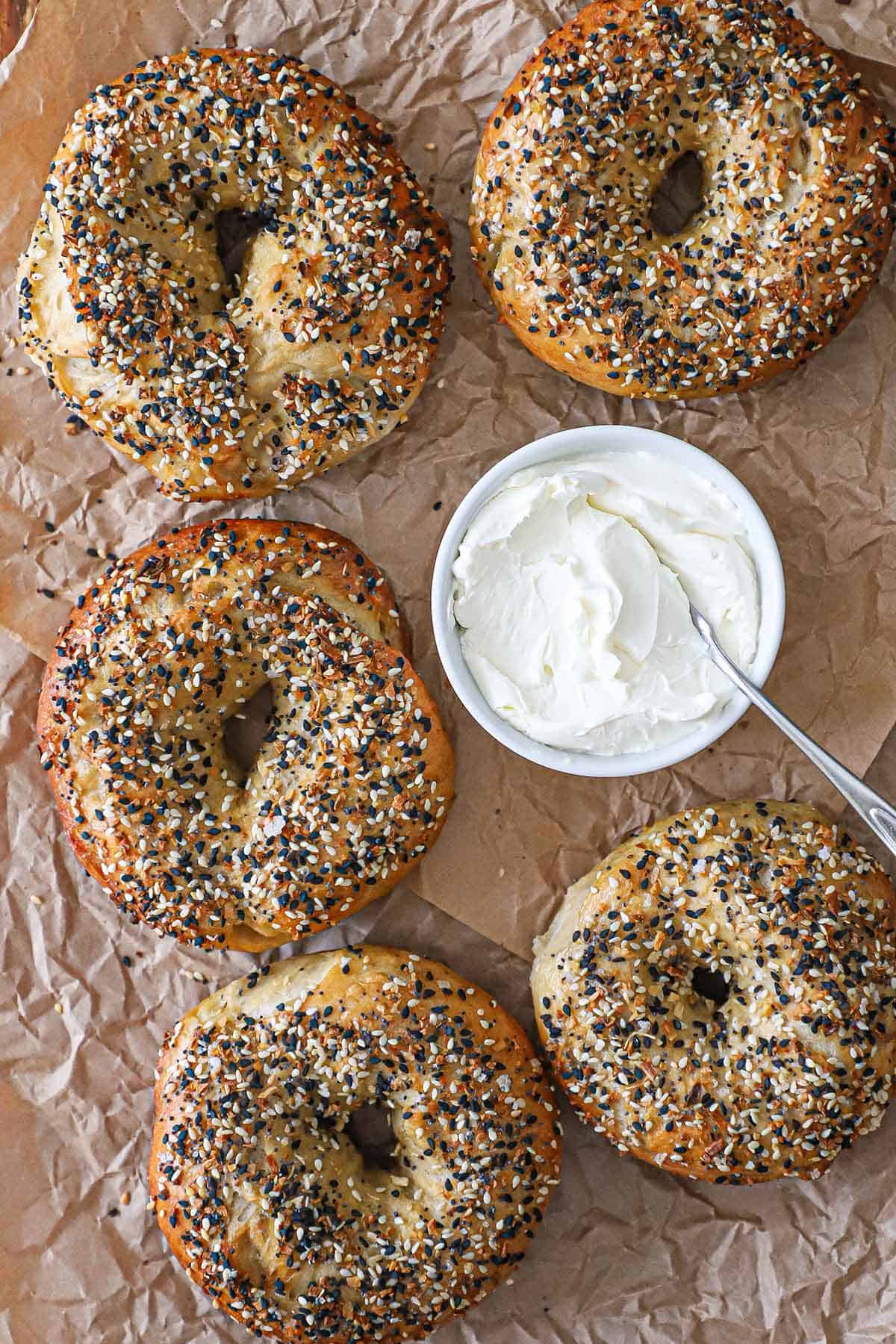 Five homemade everything bagels on a wrinkled brown piece of wax paper with a bowl of cream cheese in the middle.