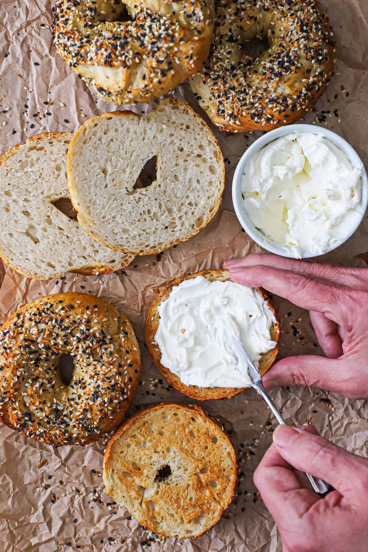 A person spreading cream cheese over the surface of a halved homemade everything bagel on a piece of brown paper next to other bagels and a bowl of cream cheese.
