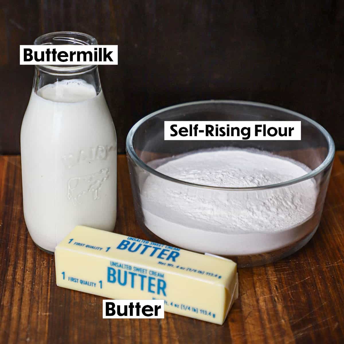 A small glass milk jug filled with buttermilk, and small glass bowl filled with self-rising flour, and a stick of unsalted butter with the wrapping still on it all sitting on a wooden cutting board.