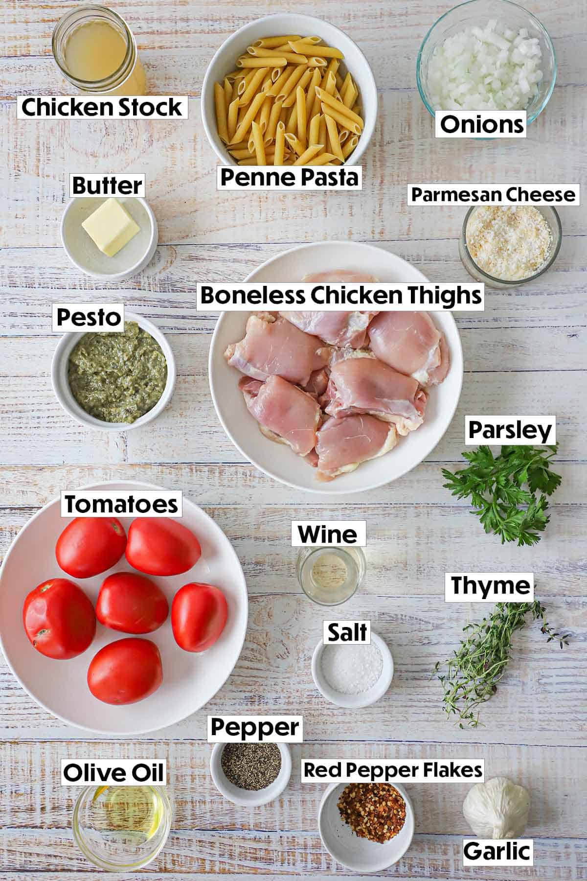 An arrangement of ingredients for pasta with roasted tomatoes and pesto chicken on a white background including a bowls of uncooked chicken thighs and Roma tomatoes, herbs, pasta, and seasonings. 