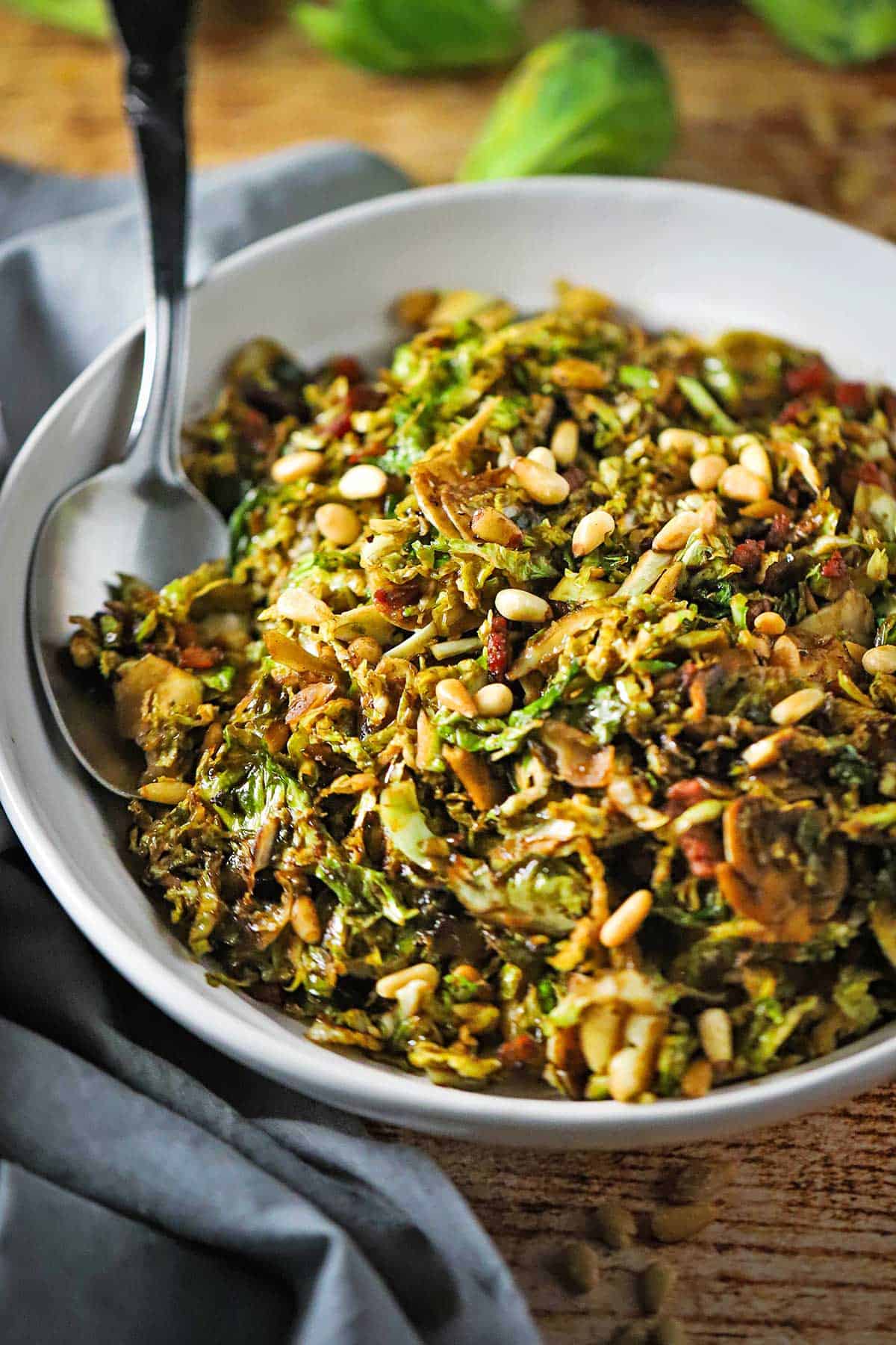 A white serving bowl filled with shaved Brussels sprouts with balsamic, pine nuts, and pancetta with a large spoon inserted into the side of the salt.