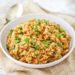 A shallow white ceramic bowl filled with a serving of chicken fried rice topped with chopped scallions and a serving spoon nearby.