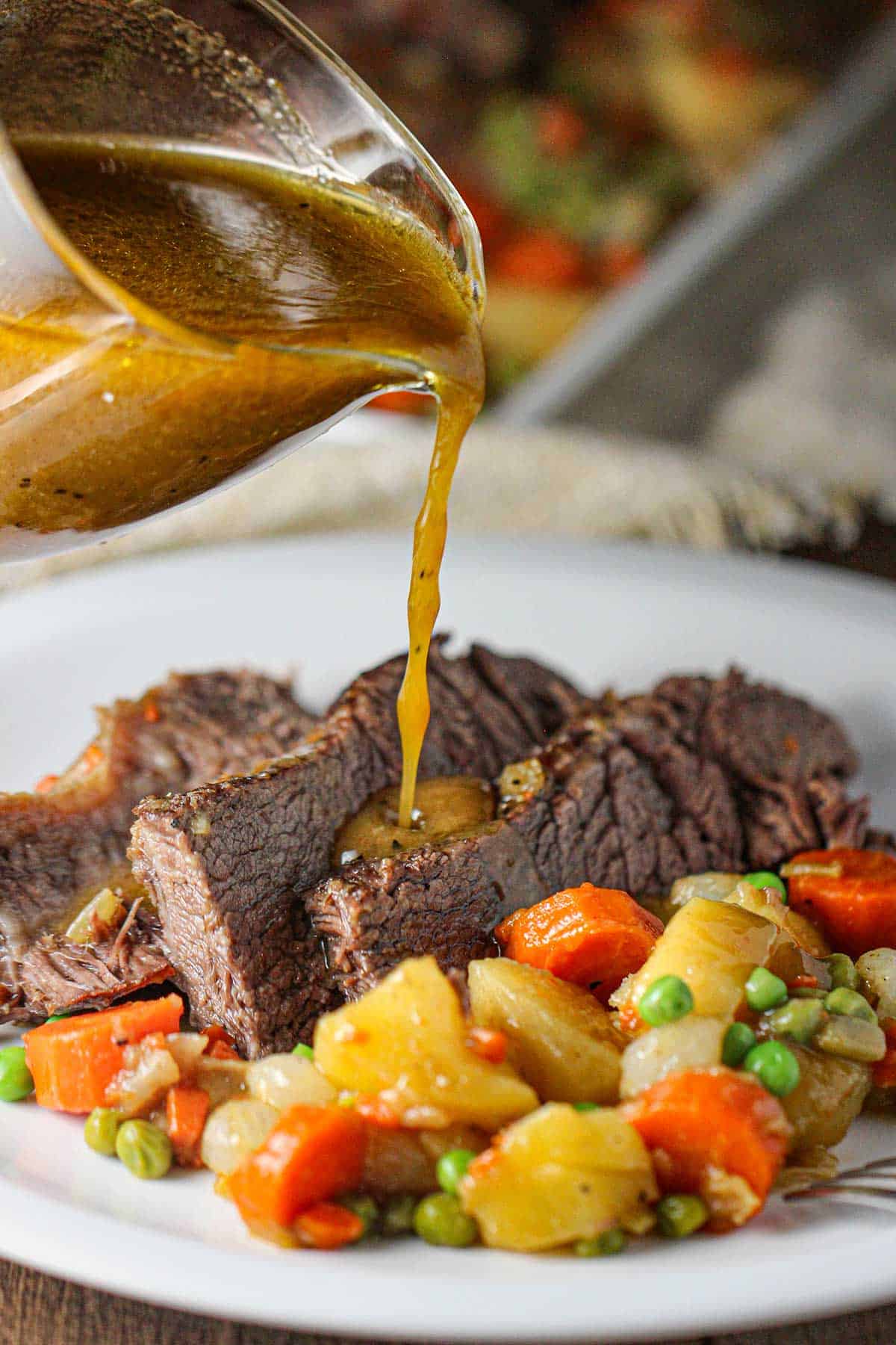 A person pouring brown gravy from a glass gravy boat onto a plate filled with three strips of classic pot roast sitting next to braised vegetables.