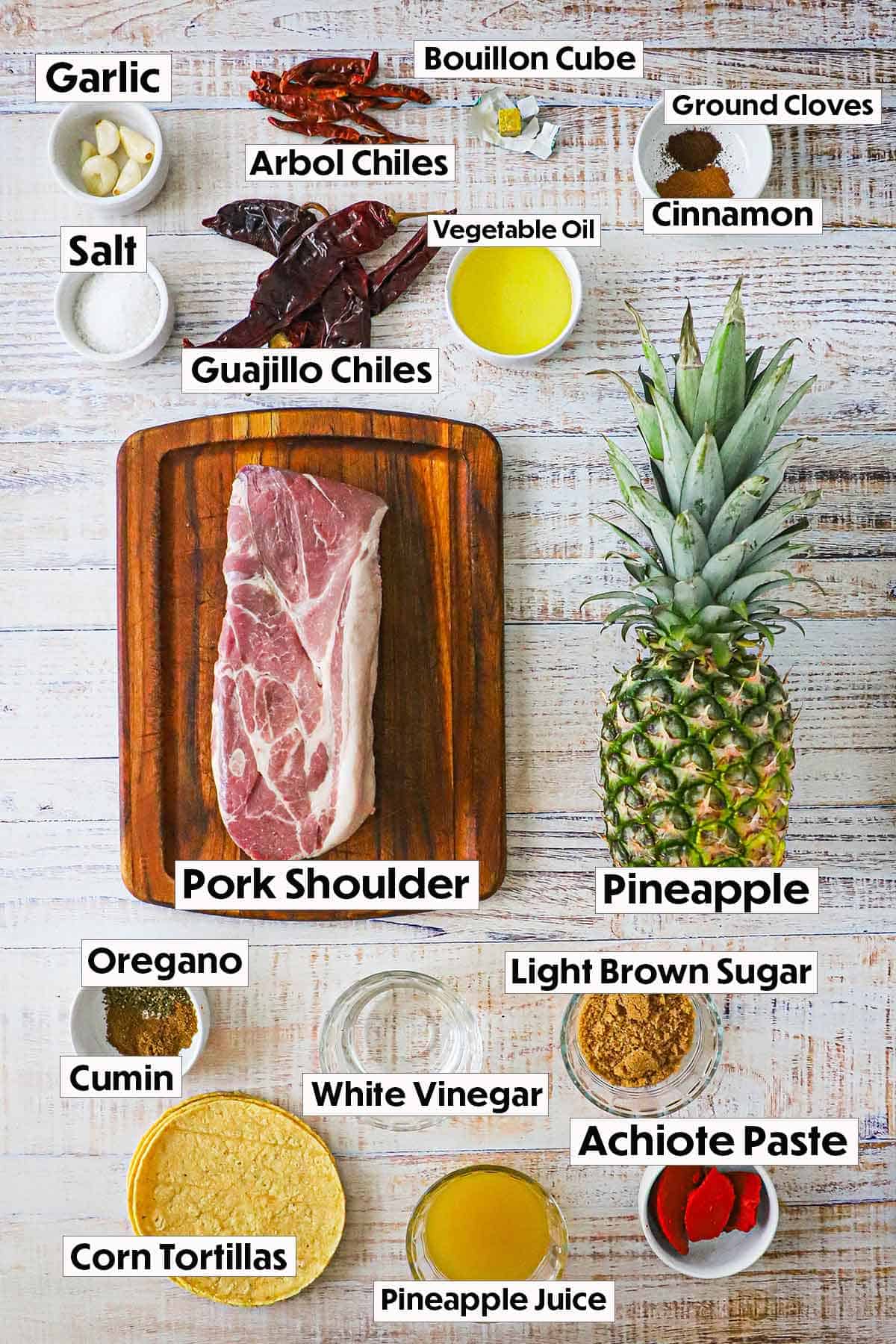 An arrangement of ingredients for grilled tacos al pastor on a white background including dried chiles, spices, pineapple, pork shoulder, sugar, vinegar, and corn tortillas.