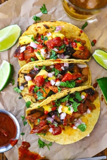 An arrangement of ingredients for grilled tacos al pastor on a white background including dried chiles, spices, pineapple, pork shoulder, sugar, vinegar, and corn tortillas.