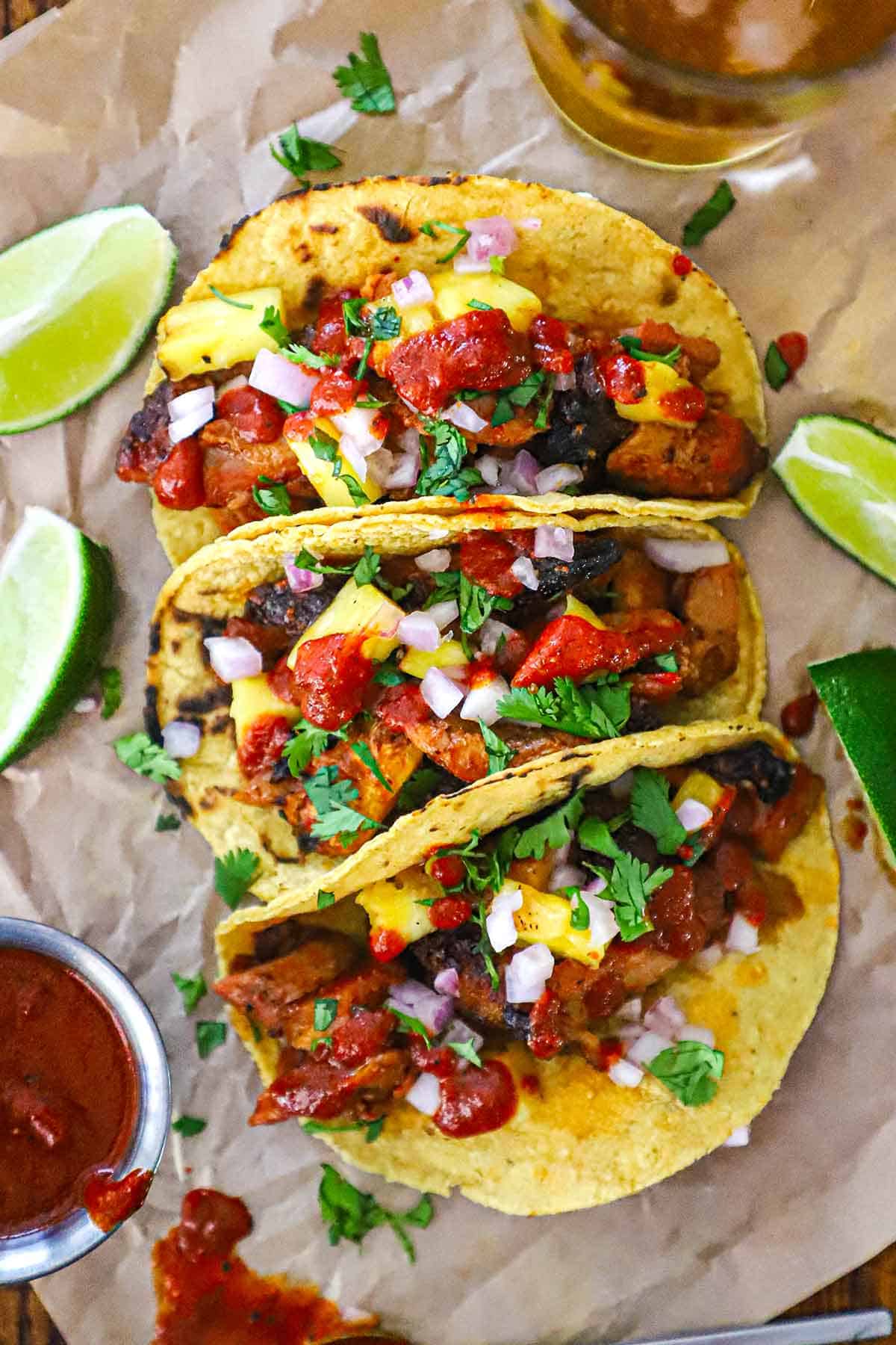 An arrangement of ingredients for grilled tacos al pastor on a white background including dried chiles, spices, pineapple, pork shoulder, sugar, vinegar, and corn tortillas.