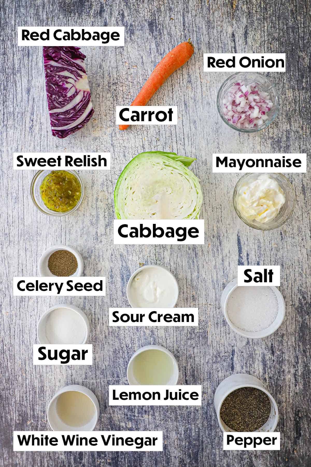 An arrangement of ingredients for diner-style coleslaw on a grey wooden background including cabbage, carrot, onion, relish, mayonnaise, sugar, sour cream, and seasonings.