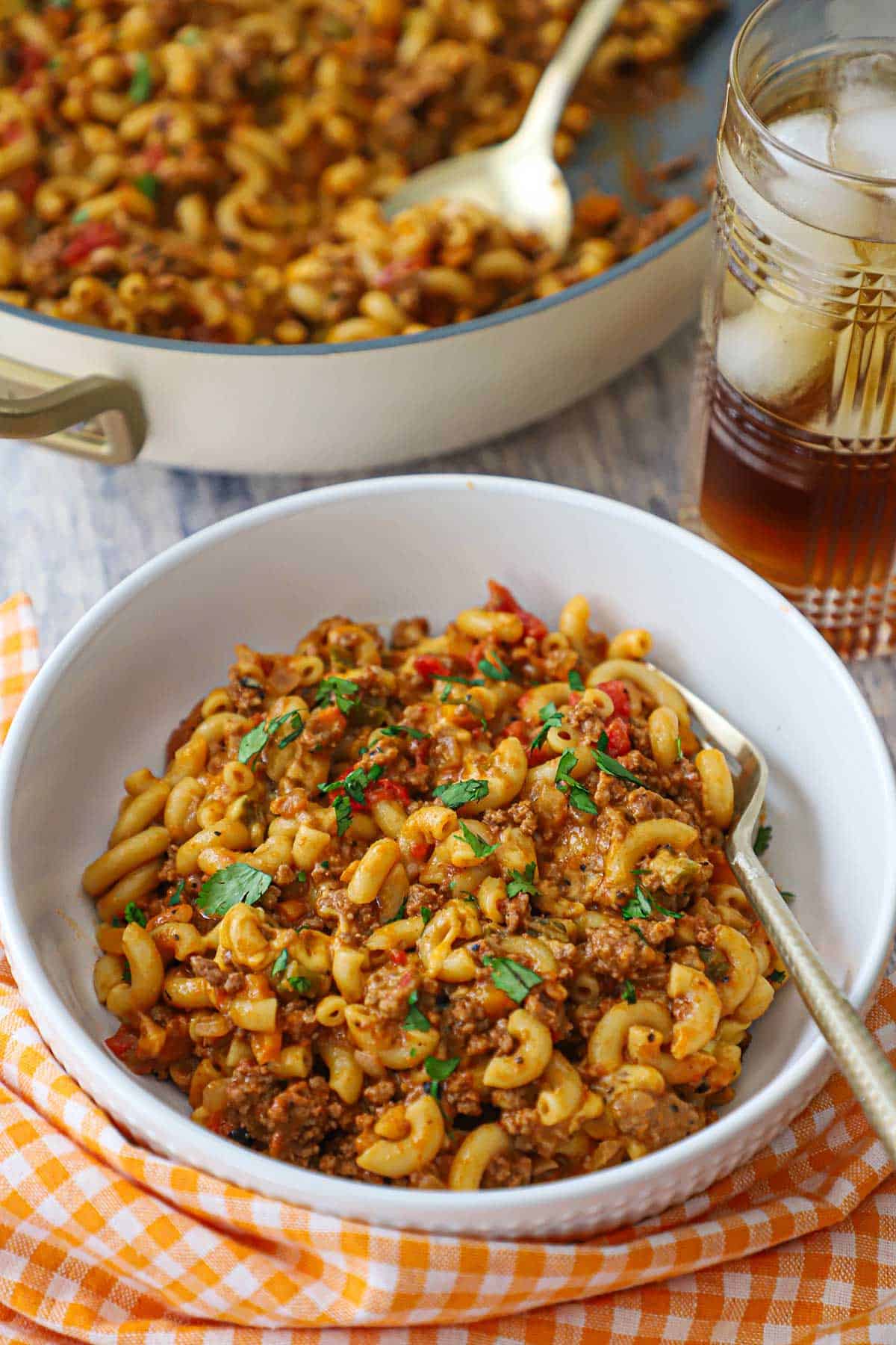 A bowl of One-Pot Chili Mac sitting net to an orange checkered napkin with a glass of iced tea and a skillet of the chili mac in the background.