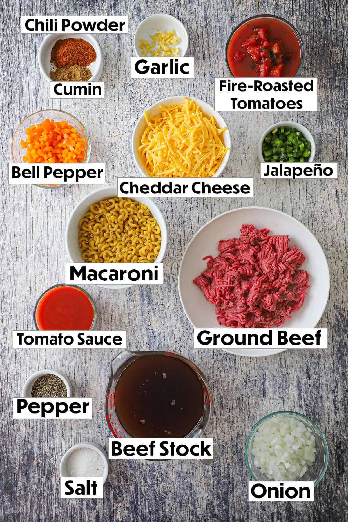 An arrangement of ingredients for chili mac on a wooden background including ground beef, seasonings, tomatoes, macaroni, peppers, and onions.