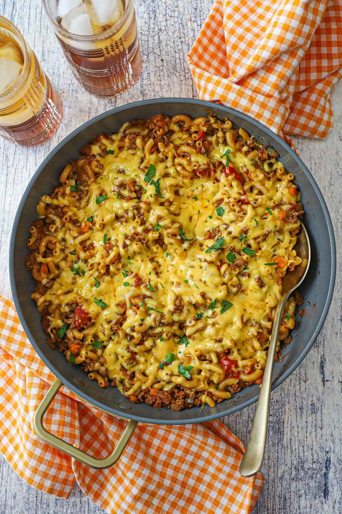 A skillet filled with one-pot chili mac with a layer of melted cheddar cheese over the the surface with colorful checkered napkins near the pan and two glasses of iced tea nearby.