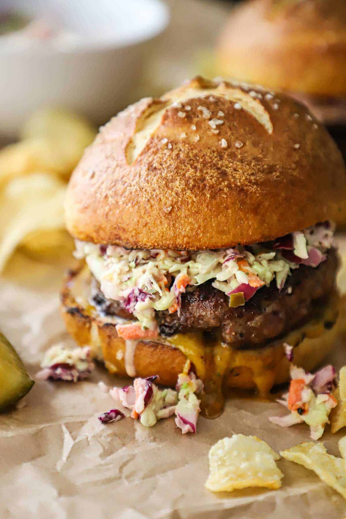 A pork burger topped with creamy coleslaw on a homemade pretzel bun with Carolina mustard sauce oozing from the underneath the patty with kettle chips scattered around the burger.