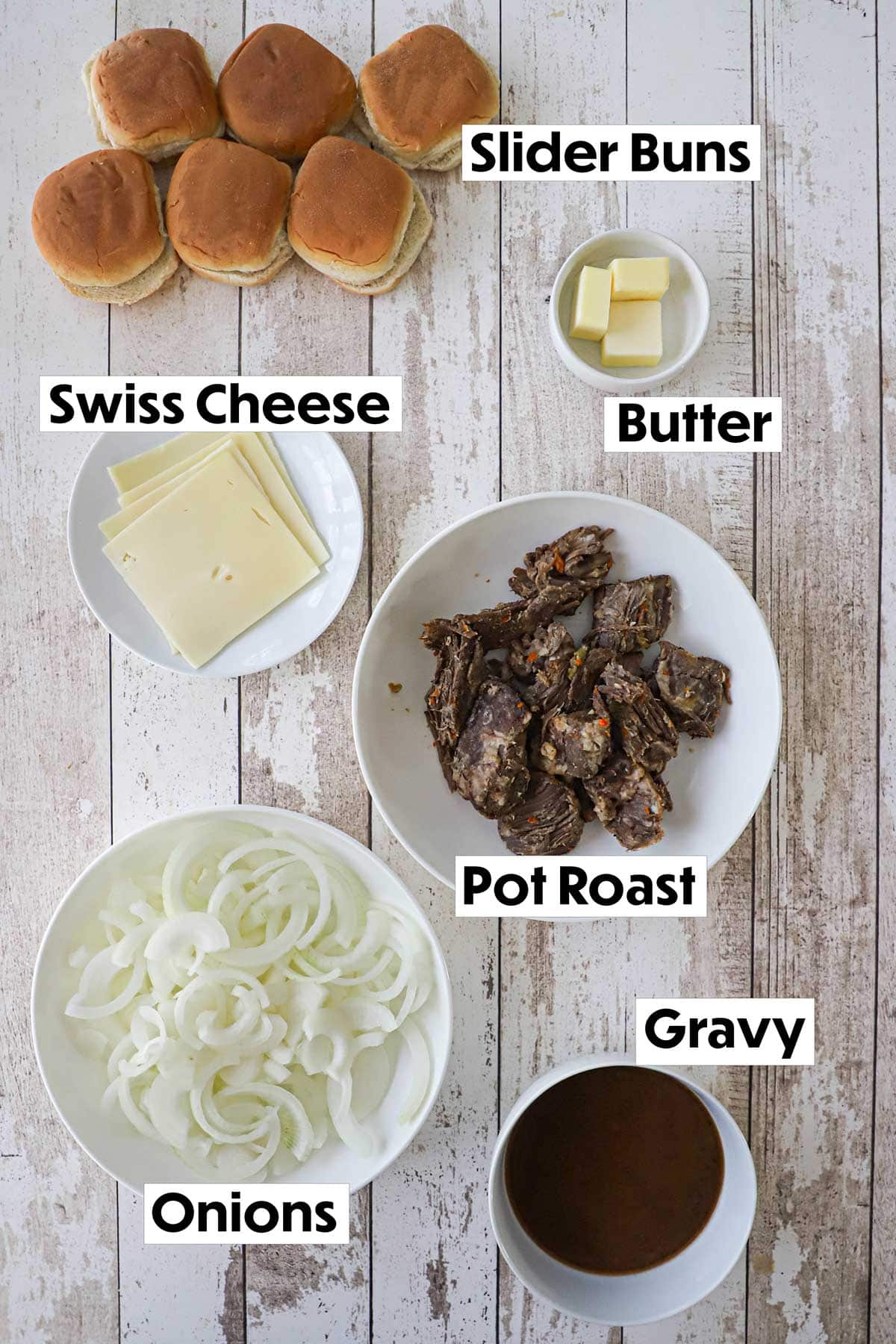 An arrangement of ingredients for pot roast sliders on a white background including slider buns, Swiss cheese slices, butter, cooked pot roast, slices onions, and a bowl of brown gravy.