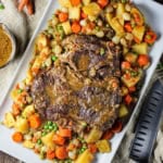 A fully cooked classic pot roast on a large rectangular white platter surrounded by cooked potatoes, carrots, and peas.