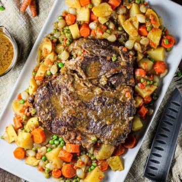 A fully cooked classic pot roast on a large rectangular white platter surrounded by cooked potatoes, carrots, and peas.