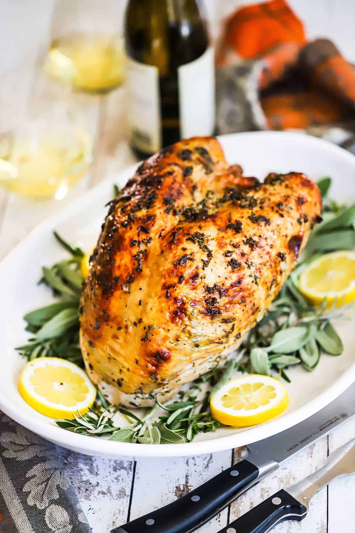 A Roast Turkey Breast with Herbs resting on a white platter garnished fresh sage leaves and lemon wedges and a large carving knife nearby.