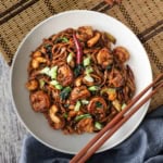 A white bowl filled with shrimp lo mein with cashews garnished with chopped scallions on top and two brown chopsticks resting side by side on the edge of the bowl.