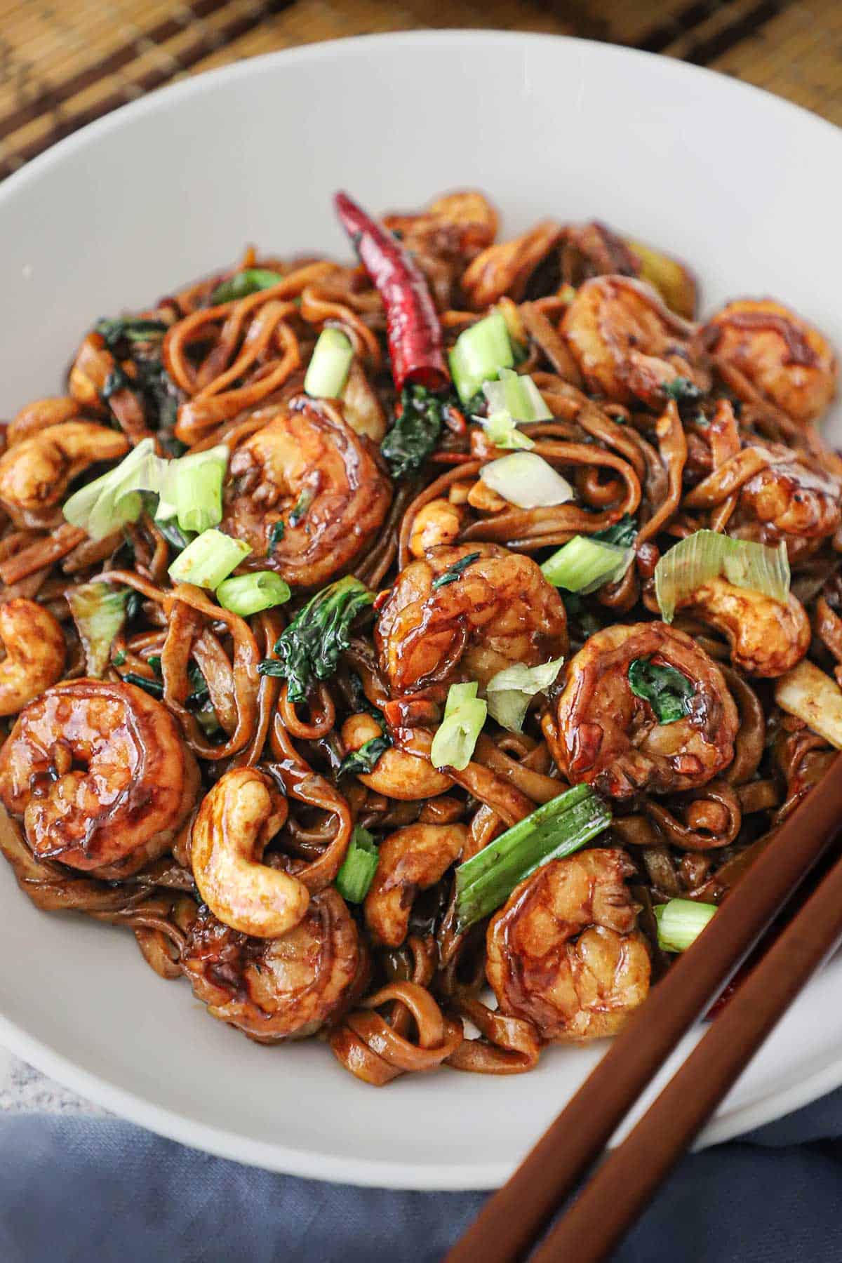 A white bowl filled with shrimp lo mein with cashews garnished with chopped scallions on top and two brown chopsticks resting side by side on the edge of the bowl.