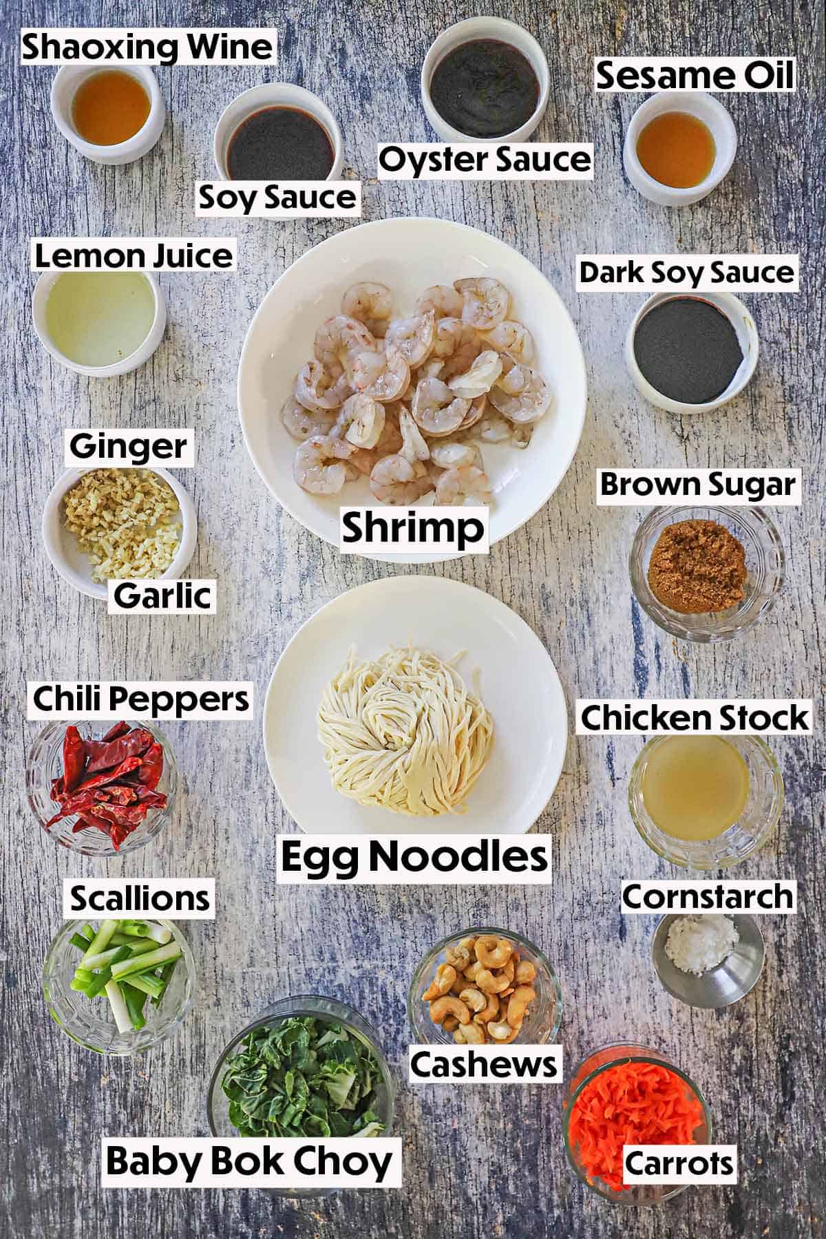 An arrangement of ingredients on a grey wooden background including uncooked shrimp, egg noodles, cashews, bok choy, carrots, chili peppers, ginger, garlic, and various bowls filled with ingredients to make the sauce.