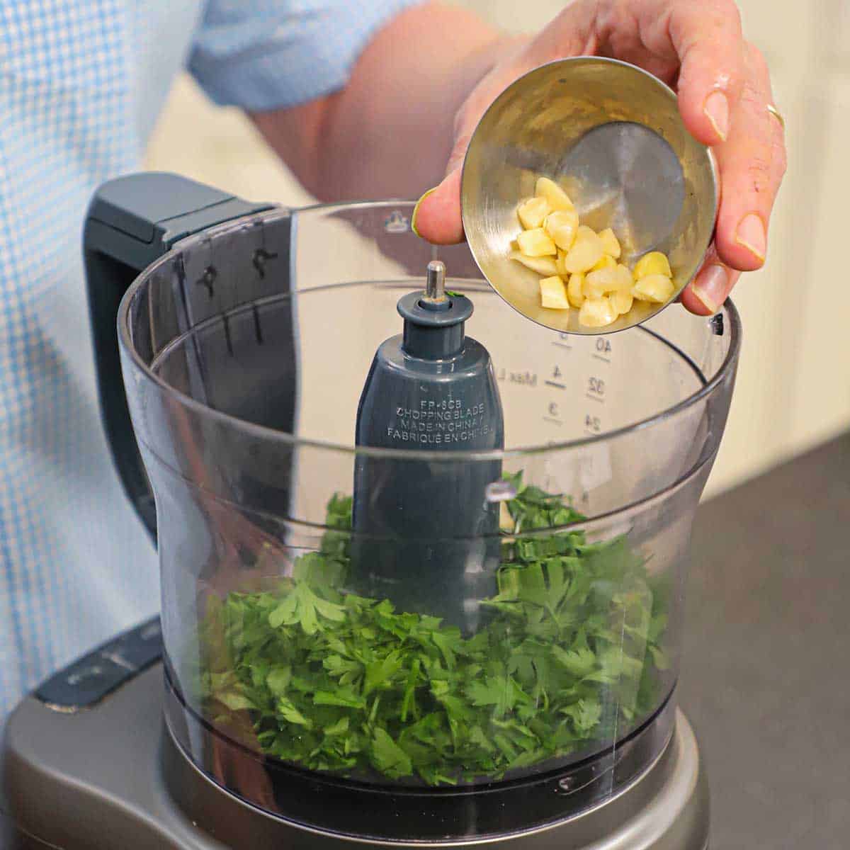 A person dumping roughly chopped garlic cloves from a small metal bowl into a food processor filled with chopped fresh Italian parsley.