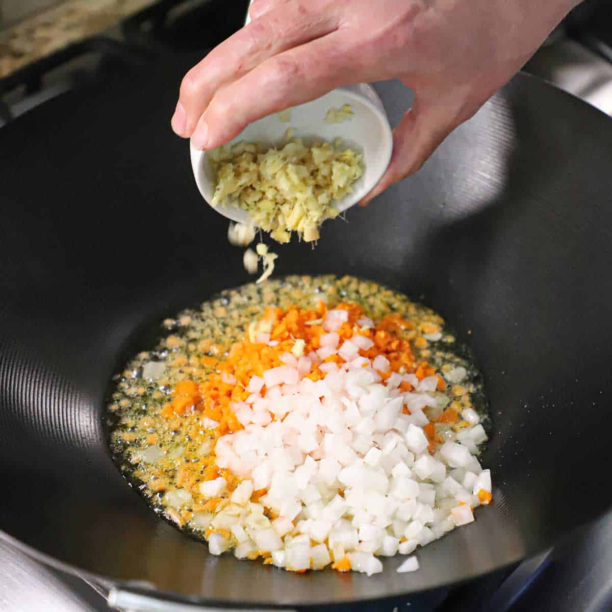 A person dumping a small bowl of chopped ginger into a wok filled with chopped carrots, garlic, and onion simmering in oil.