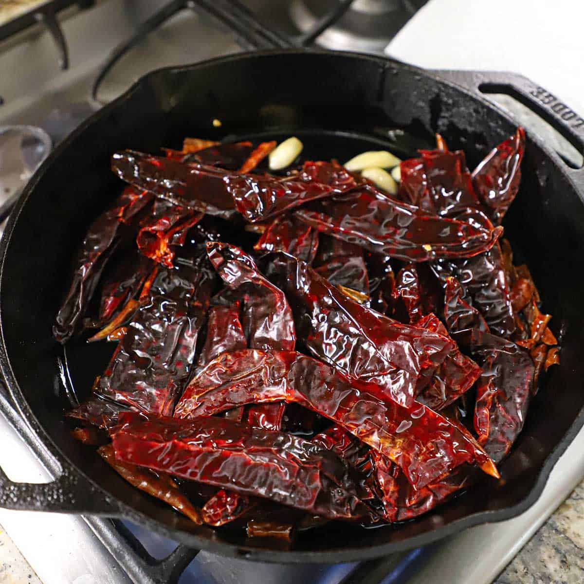 A large cast-iron skillet filled with dried chiles and cloves of garlic simmering in vegetable oil.