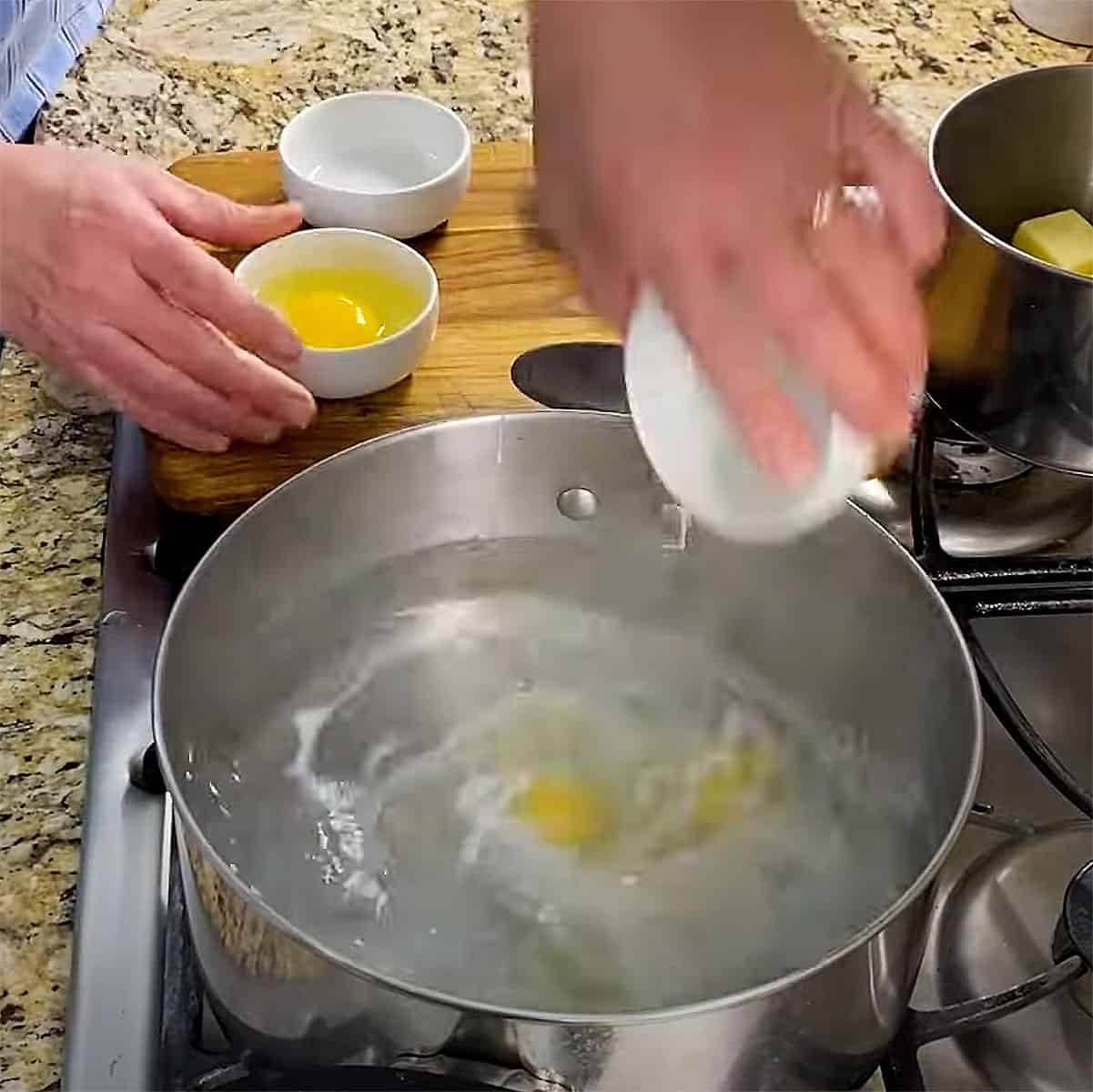 A person transferring an egg from a small white bowl into a pot of swirling boiling water in a pot on a stove.
