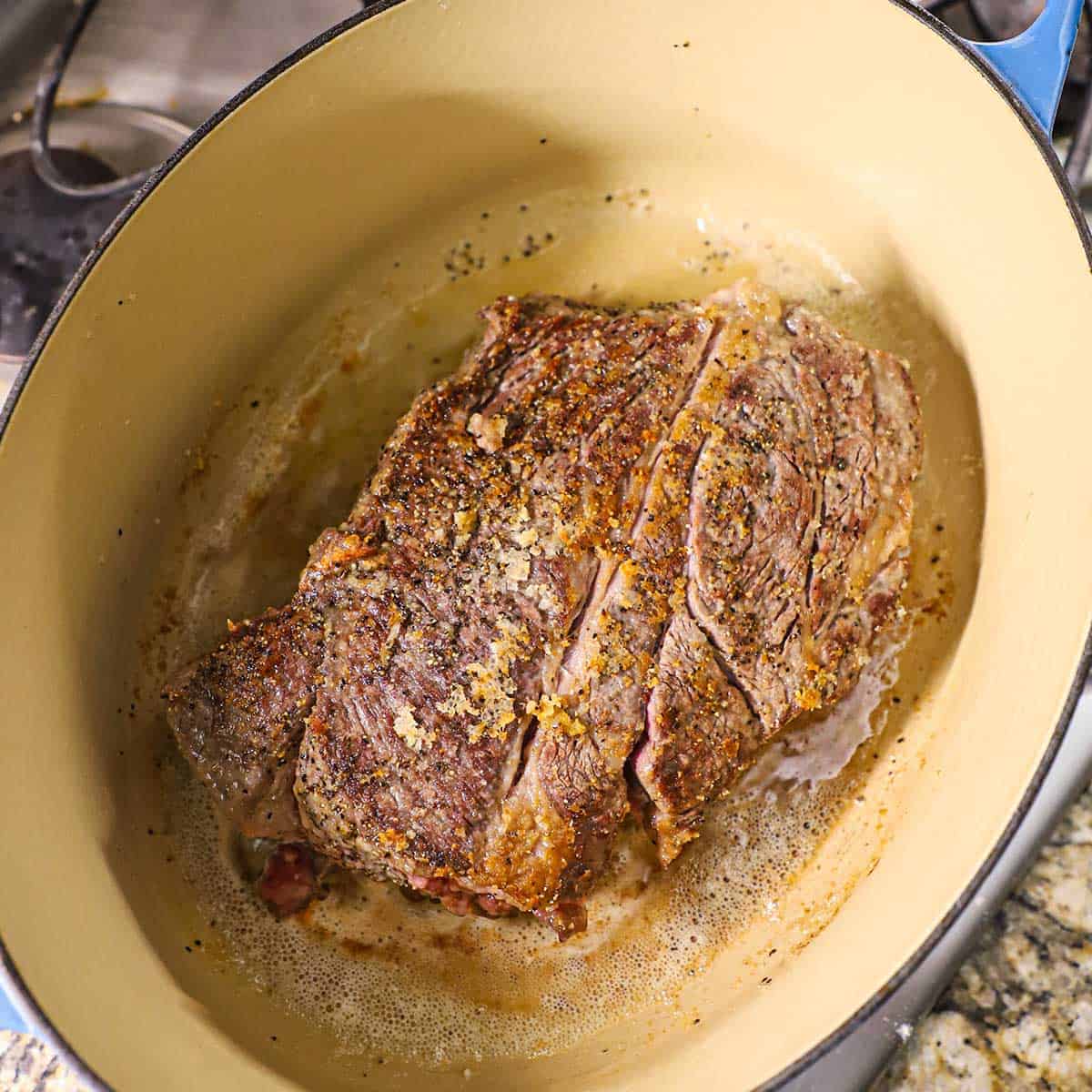 A seasoned chuck roast being seared in sizzling oil in a large oval Dutch oven on a gas stove.