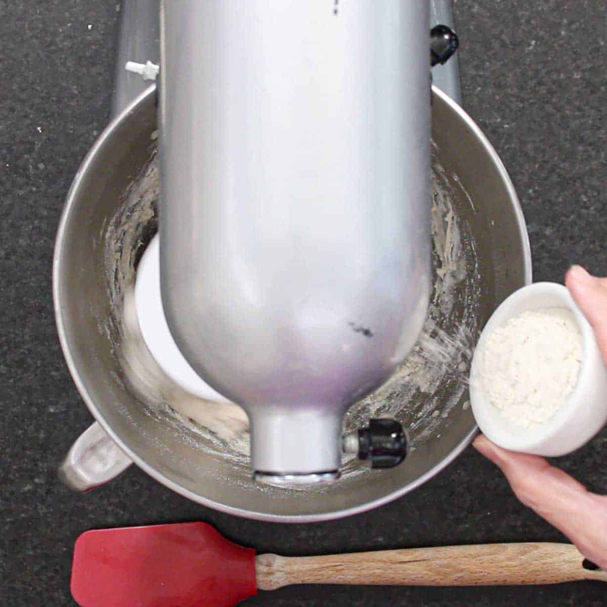 A person adding a small amount of flour from a white bowl into a stand mixer that is kneading bagel dough.