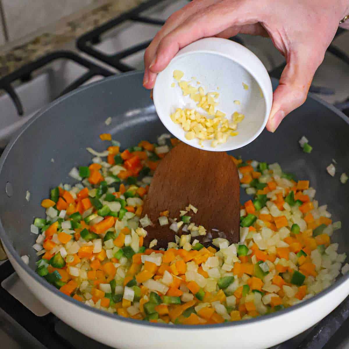 A person dumping minced garlic from a small white bowl into a skillet filled with sautéed diced onions, orange bell pepper, and jalapeño.