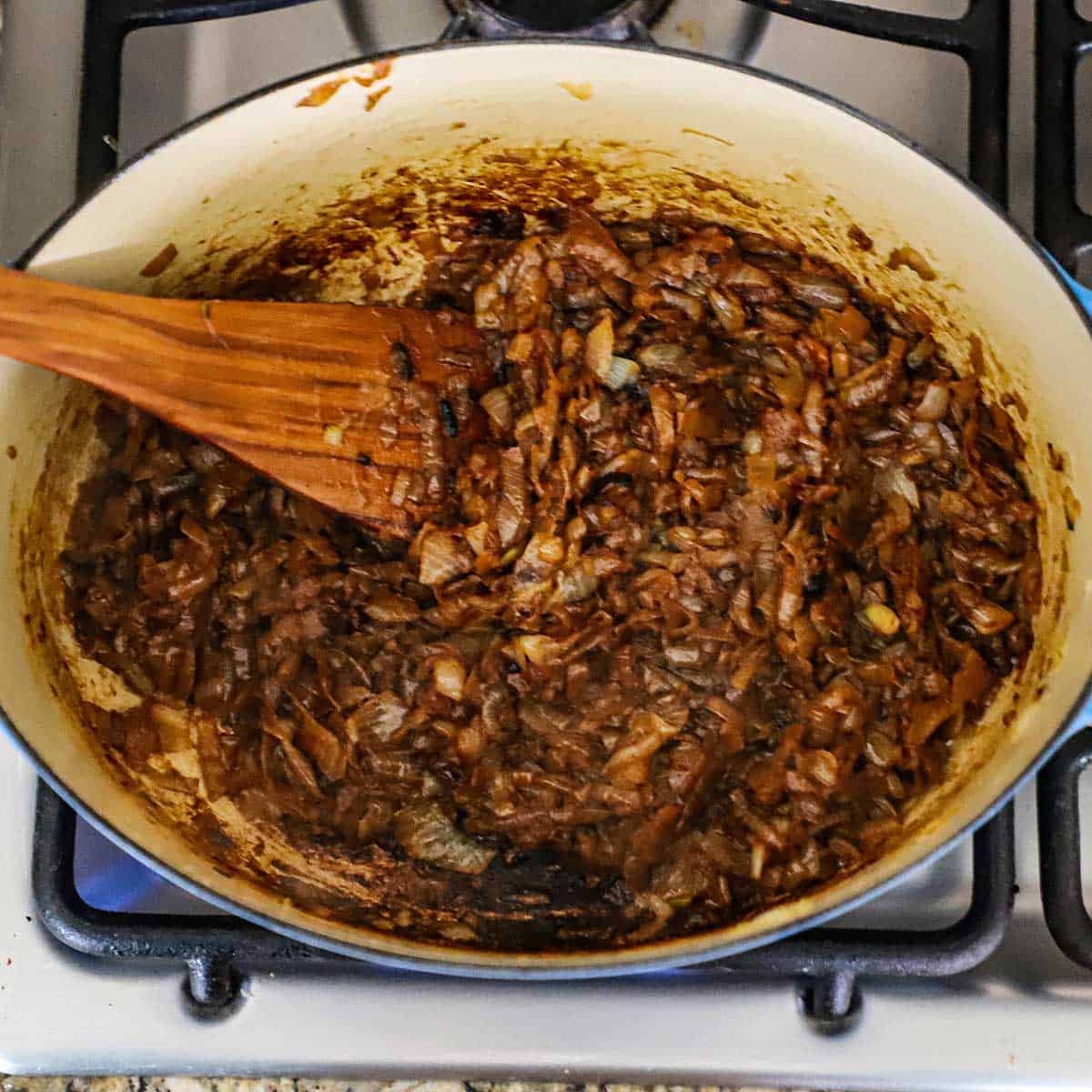 A large blue Dutch oven filled with dark caramelized onions with a wooden spatula stuck into the side.