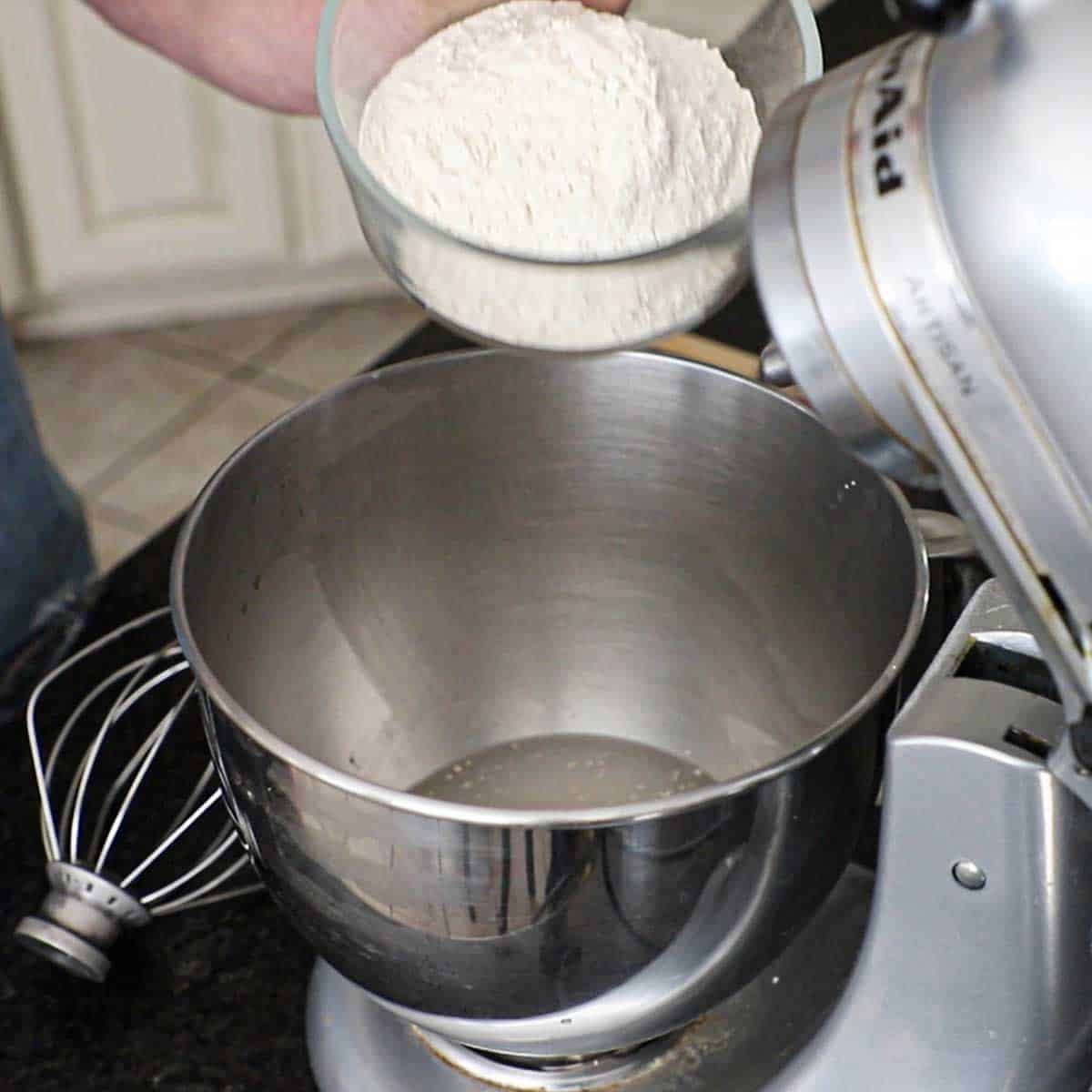 A person holding a glass bowl filled with high-gluten flour that he is about to dump into a mixer bowl that has foamy yeast water in it.