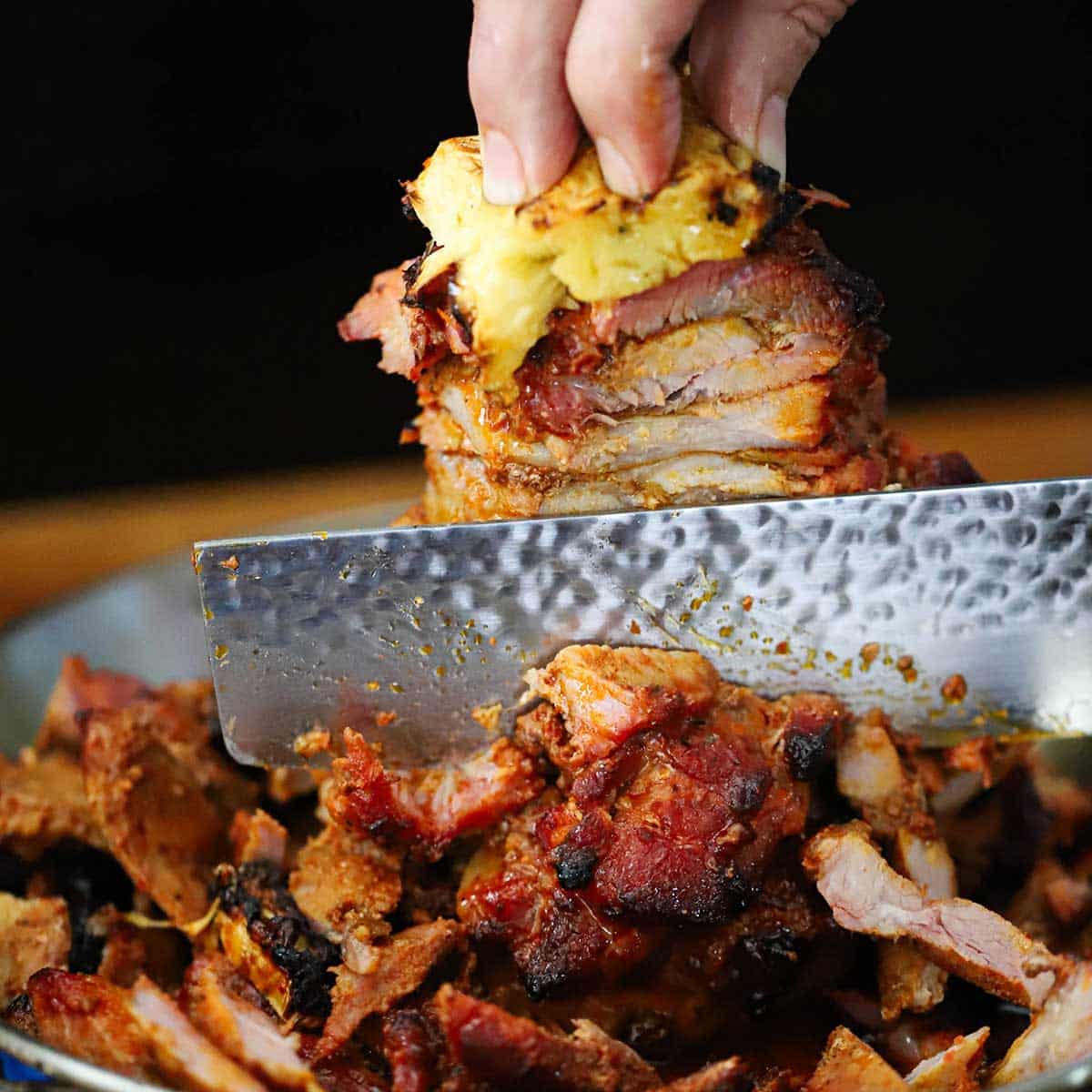 A person using a chef's knife to slice chunks of cooked marinated pork from a skewer that has a slice of pineapple on top.