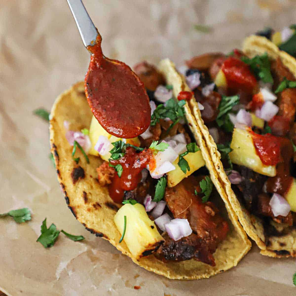 A person using a small spoon to add a layer of homemade taco sauce over the top of tacos al pastor resting on brown wax paper.
