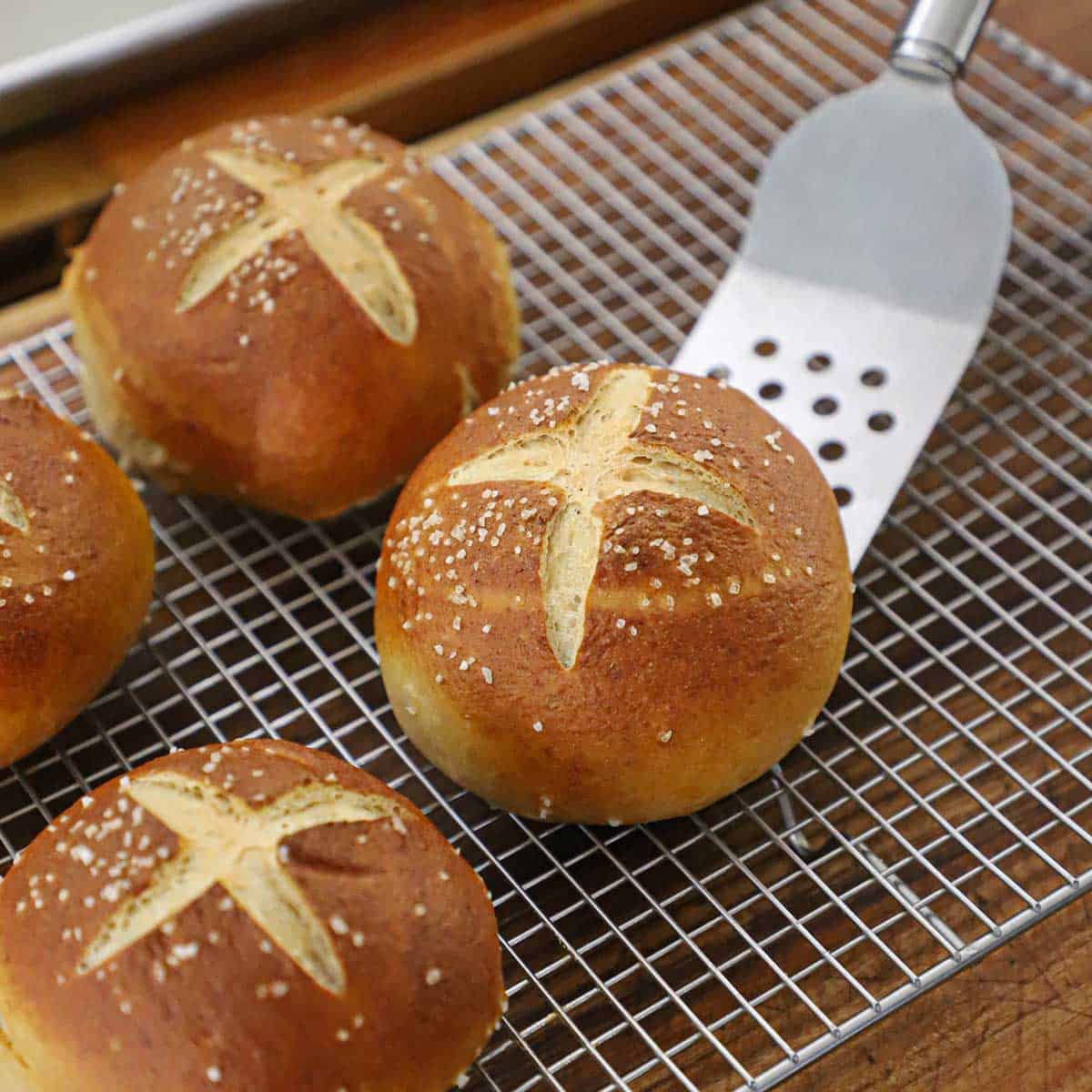 A metal spatula being used to place a freshly baked homemade pretzel bun onto a baking rack with three other pretzel buns on it.